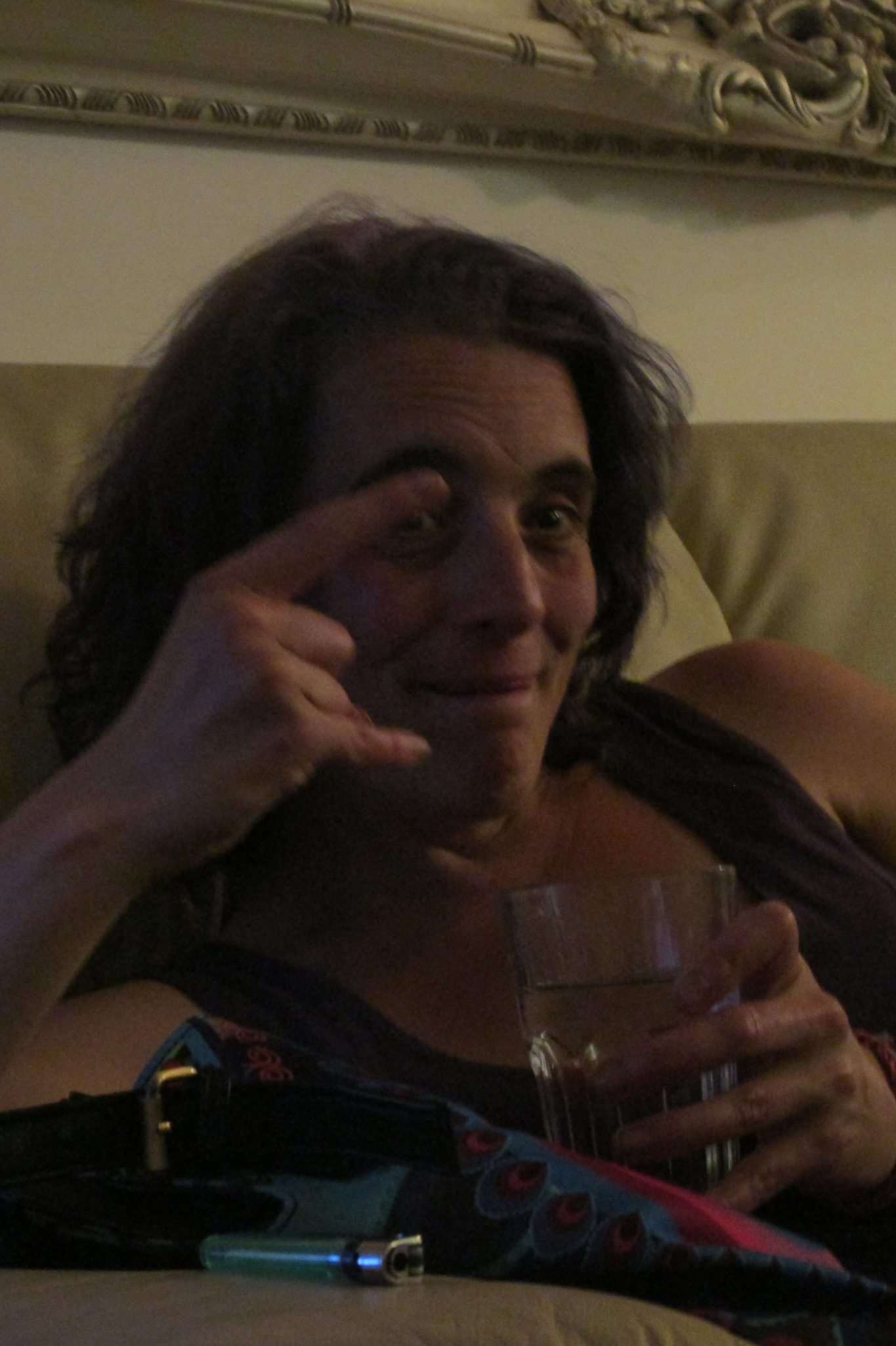 Esther with a glass of something alcoholic in her hand, her lighter to the side of her, and she's making the rock horns symbol with her hand. She is trying to smile but looks drunk.